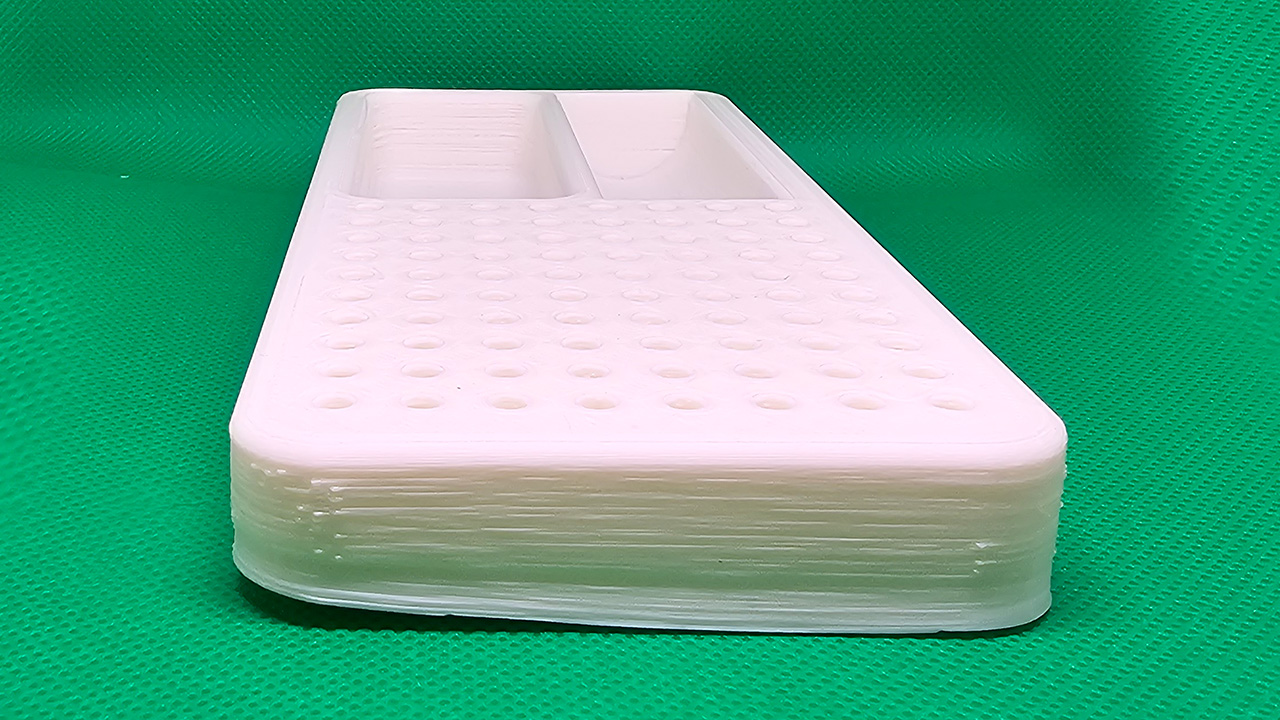 3D printed white plastic organizer tray with two compartments, a perforated base, and visible layer lines, sitting on a green surface used with blue tape.