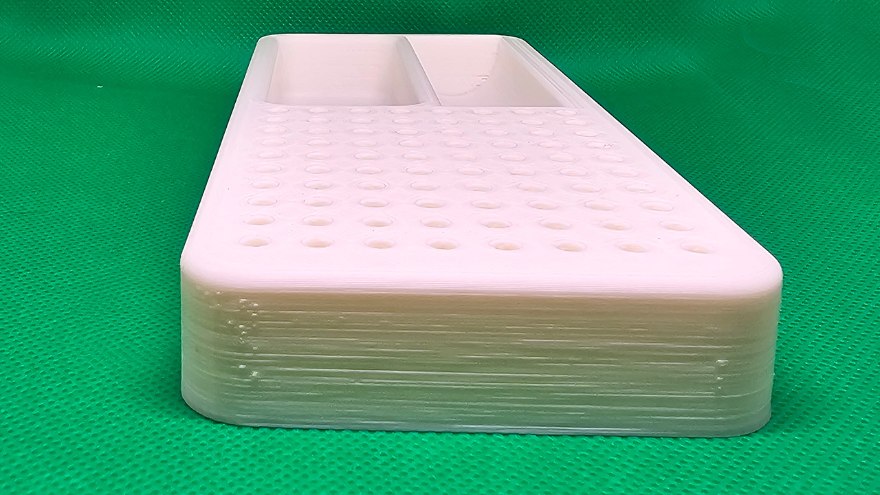 A 3D-printed white plastic organizer tray with two compartments and rows of small holes on a green grid surface, with a PEI bed. Now to dial in and make the layers look better.