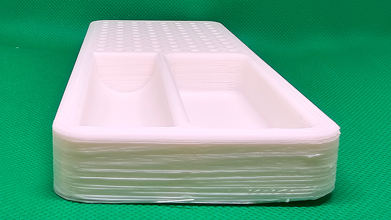 3D printed white plastic tray with two compartments, resting on a green surface. Visible layer lines show the additive manufacturing process with the default Ender 3 bed.